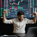 A person in a white shirt raises their fists in celebration in front of a laptop, with a large screen showing financial graphs and charts in the background.