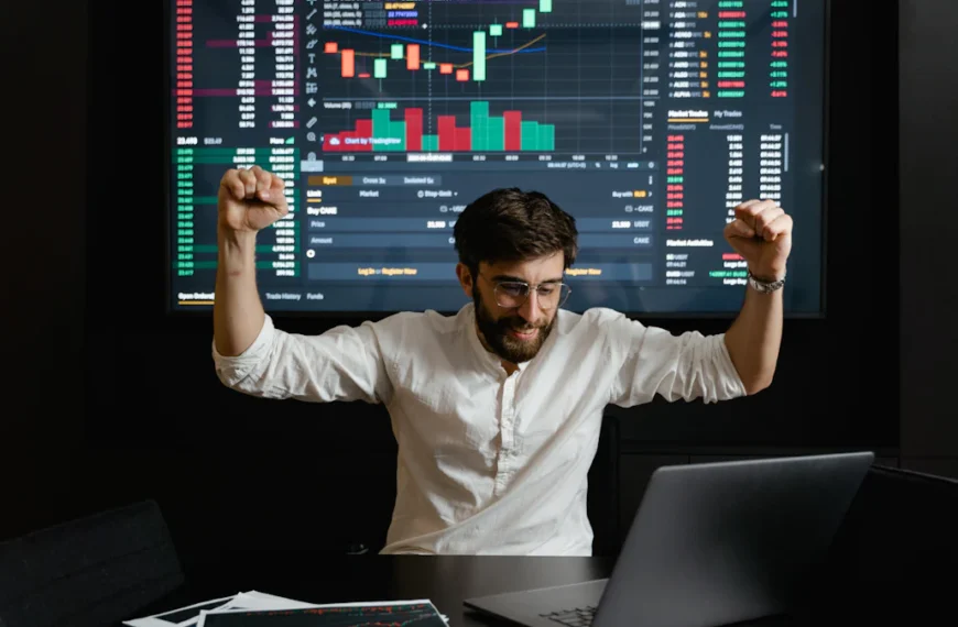 A person in a white shirt raises their fists in celebration in front of a laptop, with a large screen showing financial graphs and charts in the background.