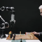 A man with a beard plays chess against an industrial robotic arm on a small table.