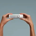 Two hands hold a rectangular medical device displaying colored waveforms and readings on a screen, against a plain gray background.