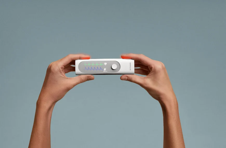 Two hands hold a rectangular medical device displaying colored waveforms and readings on a screen, against a plain gray background.