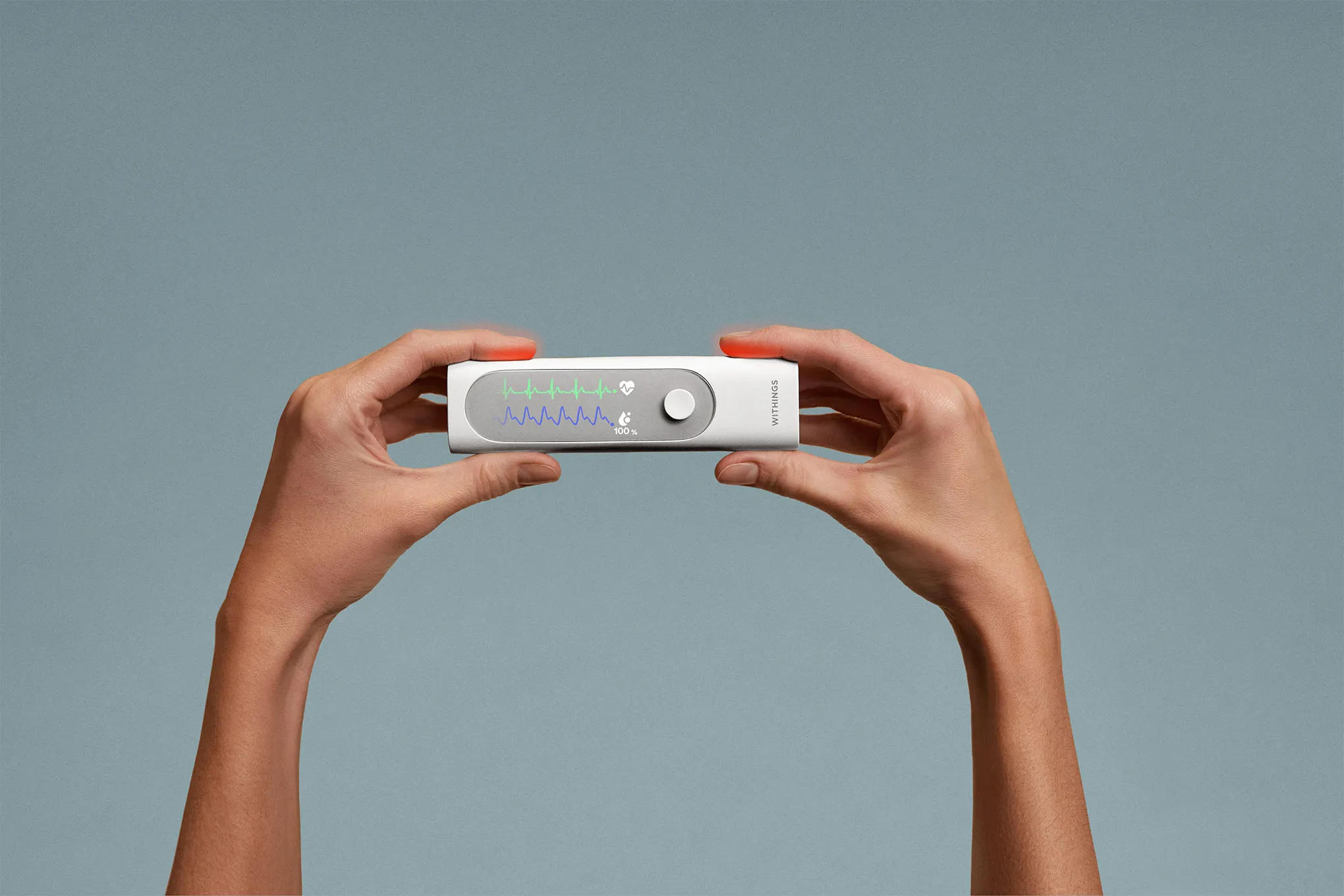 Two hands hold a rectangular medical device displaying colored waveforms and readings on a screen, against a plain gray background.